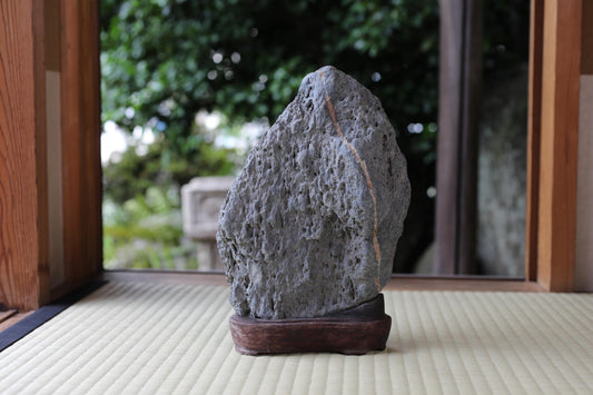 A large, gray rock sits on a wooden base in a serene garden setting, with a blurred background of trees and foliage.