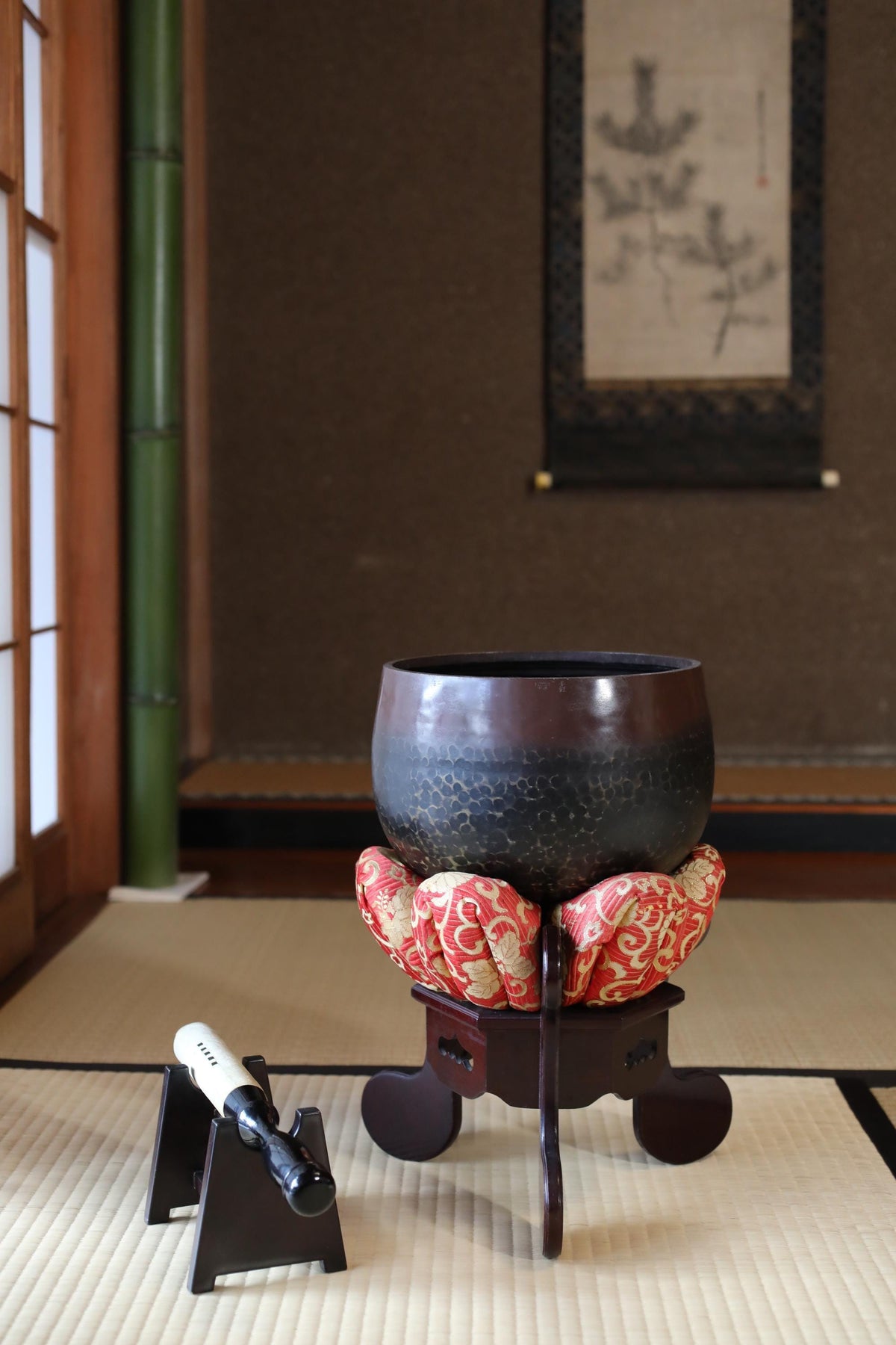 a traditional Japanese room with a large ceramic bowl on a wooden stand, a small wooden tray with a bottle, and a framed picture on the wall.