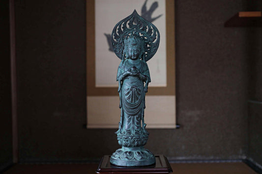 A bronze statue of a Buddhist deity, likely a Buddha, is displayed on a wooden stand in a dimly lit room.