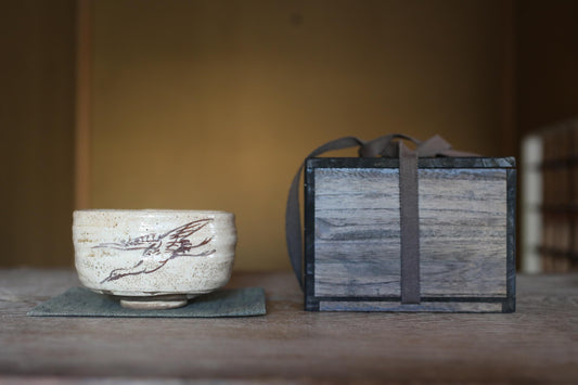 A white ceramic bowl with a bird design sits on a wooden surface next to a wooden box with a gray ribbon tied around it.