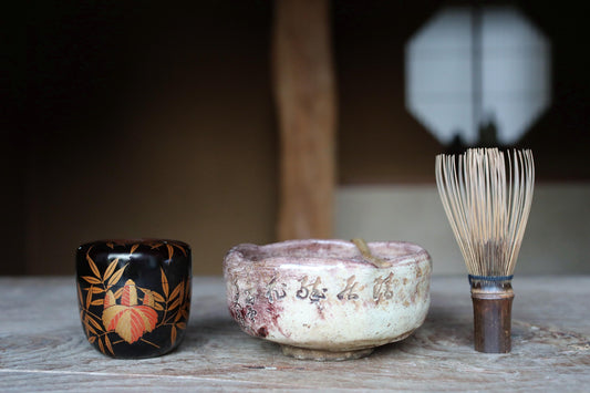a wooden table with three objects on it: a black ceramic cup with a floral design, a white ceramic bowl with Chinese characters, and a wooden whisk with a long handle.