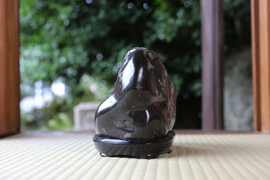 A black stone sculpture is displayed on a wooden surface, with a blurred natural background.