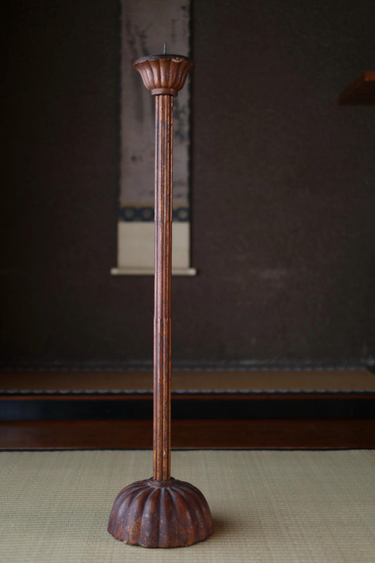 A tall, ornate wooden candle holder stands on a beige mat against a dark background.
