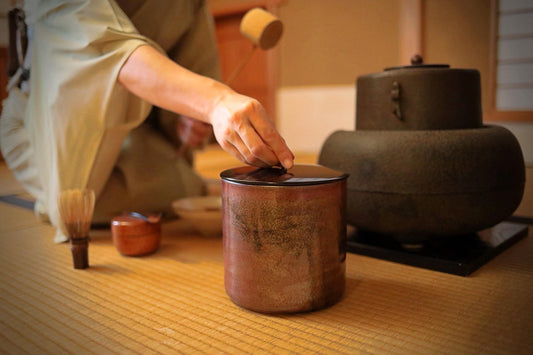 A person is carefully placing a small object into a red ceramic container on a wooden surface.