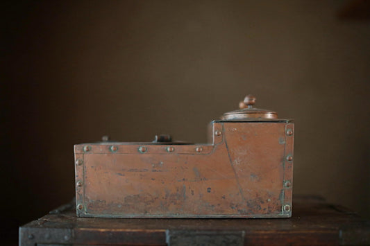 An old, weathered metal box with a lid and a small handle on top, sitting on a wooden surface against a dark background.