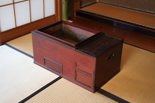 A wooden box with a lid and drawers sits on a tatami mat in a room with a sliding door and a wooden floor.