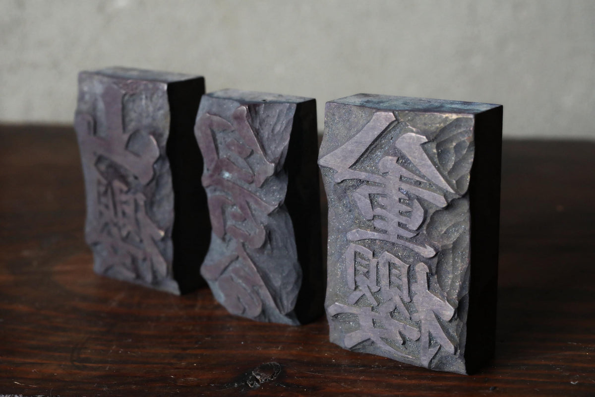 Three dark-colored rectangular blocks with carved designs, likely made of wood or stone, arranged on a wooden surface.
