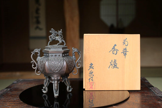 a decorative metal incense burner with an ornate lid, placed on a wooden surface next to a wooden box with Japanese characters on it.