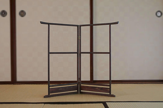 A wooden folding screen or gate with a curved top and bottom stands in the center of a room with a tatami mat floor and a traditional Japanese-style wall.