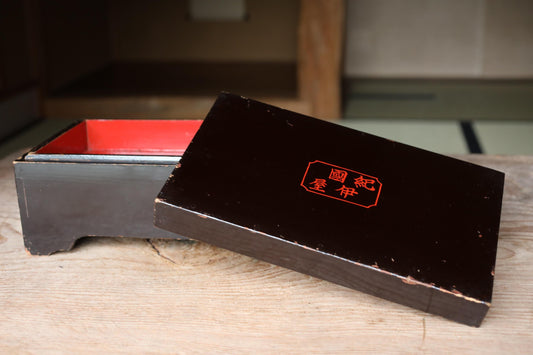 A black wooden box with a red interior, featuring an orange Chinese character on its lid, is placed on a wooden surface.