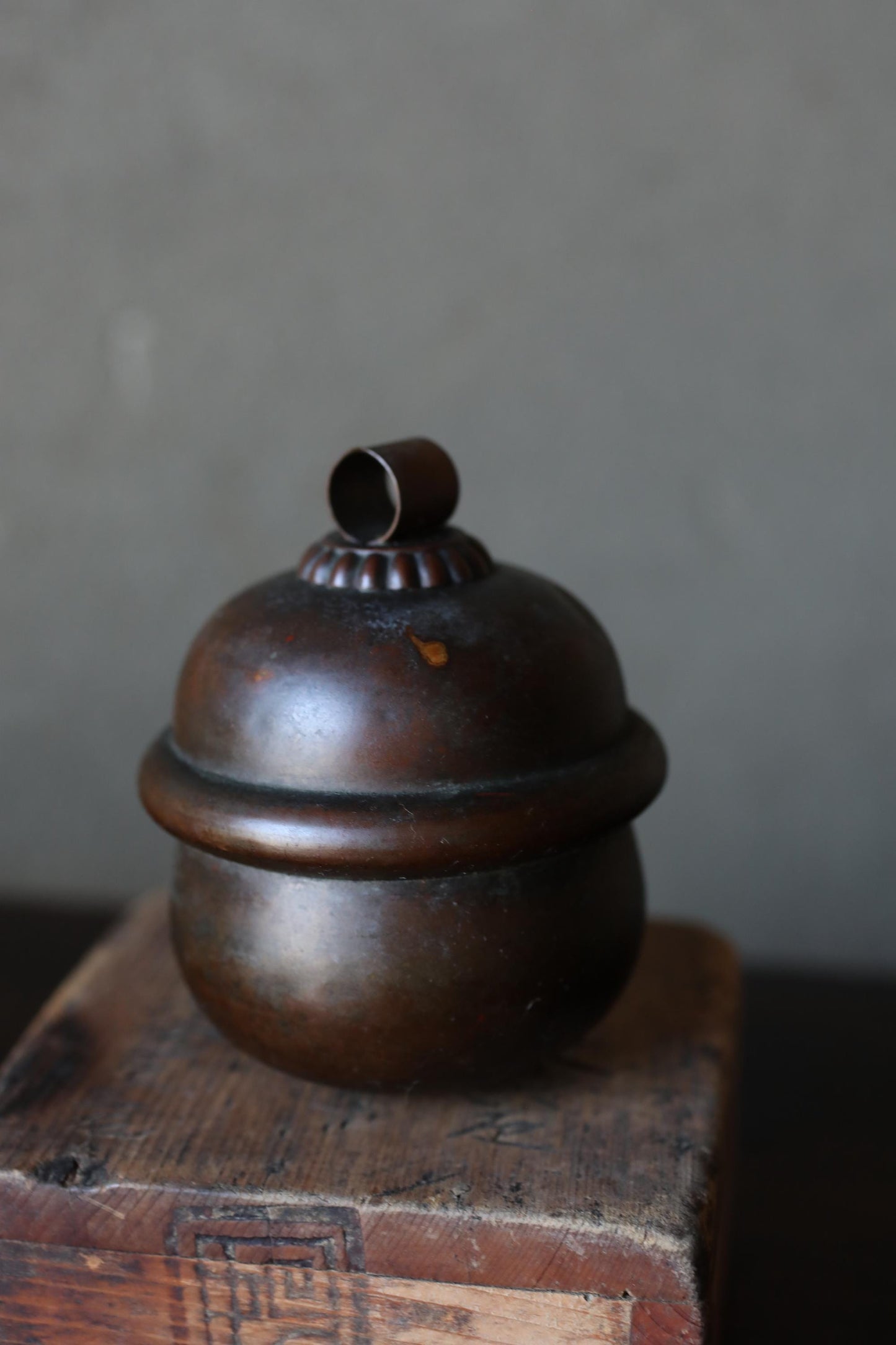 A dark brown, round metal container with a small, round finial on top, sitting on a wooden surface.