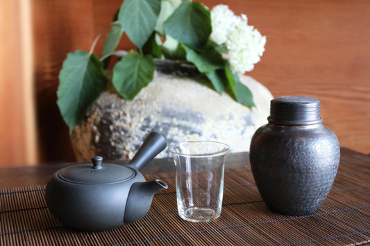 A black teapot, a glass cup, and a metal vase are arranged on a wooden table, with a vase of white flowers in the background.