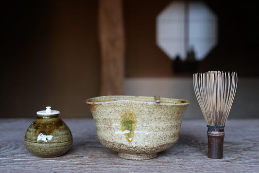 a ceramic bowl, a small ceramic jar, and a wooden whisk on a wooden surface.
