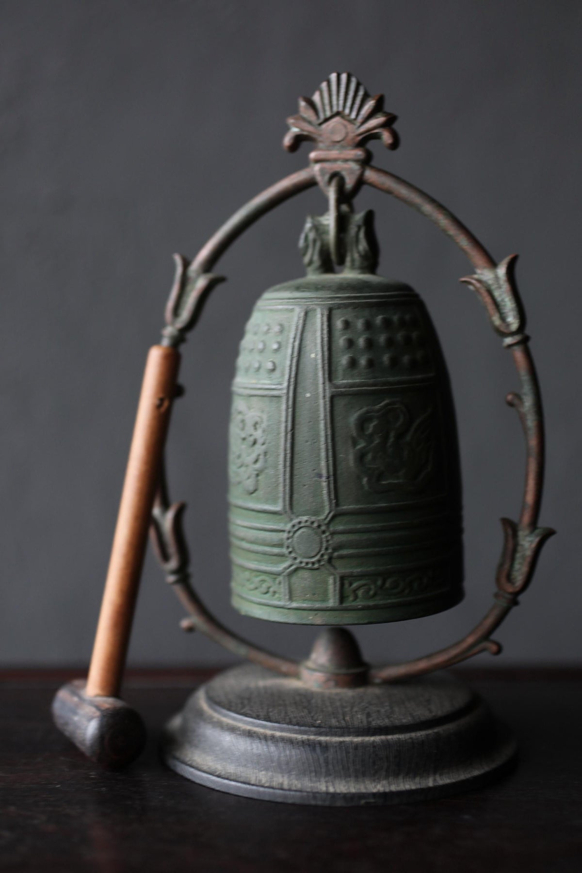 An antique green metal bell with a decorative metal frame, placed on a dark surface.