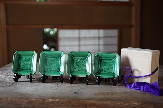 A row of four green ceramic plates with intricate designs, each placed on a wooden stand, accompanied by a purple string and a wooden box.
