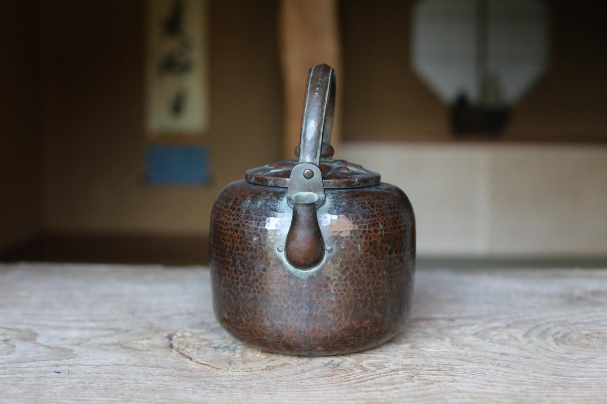 An old, copper-colored kettle with a handle and spout sits on a wooden surface, with a blurred background suggesting an indoor setting.