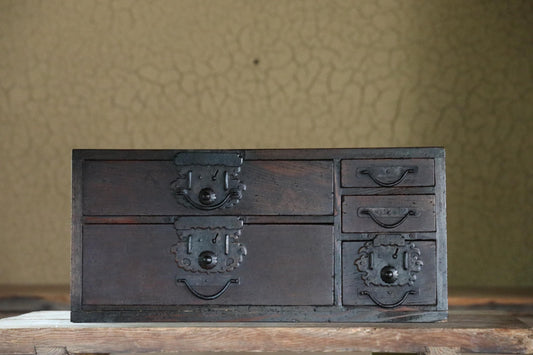 A dark wooden chest with multiple drawers and ornate metal handles is displayed on a wooden surface against a textured wall.