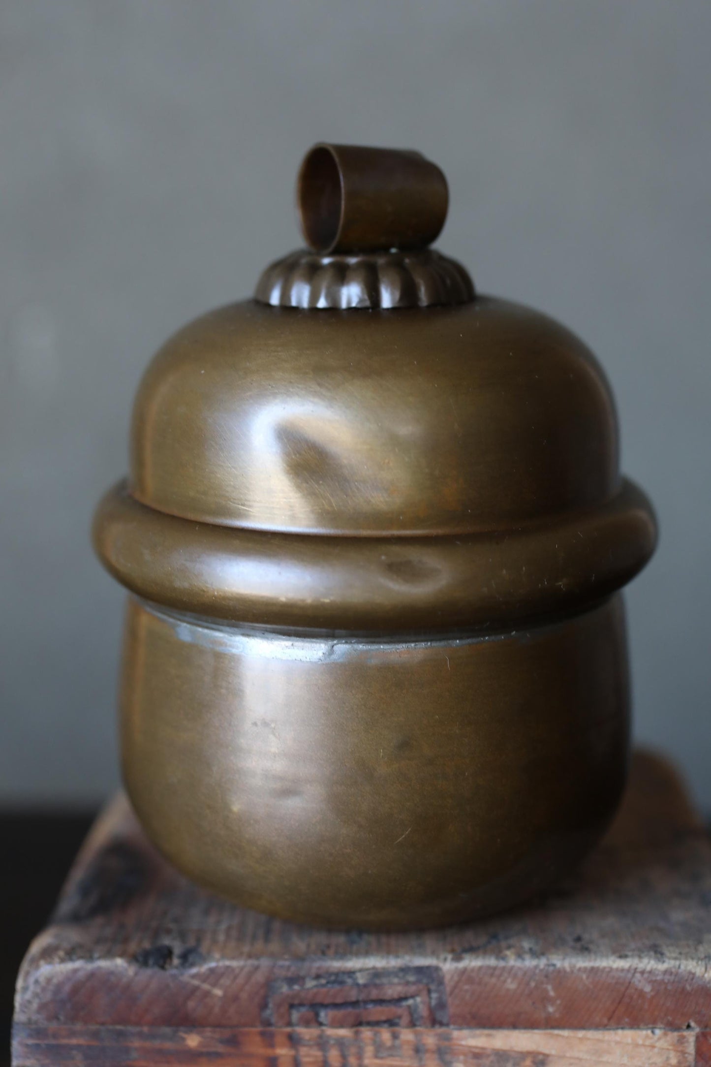 A shiny, brass-colored metal container with a lid and a small metal piece on top, sitting on a wooden surface.