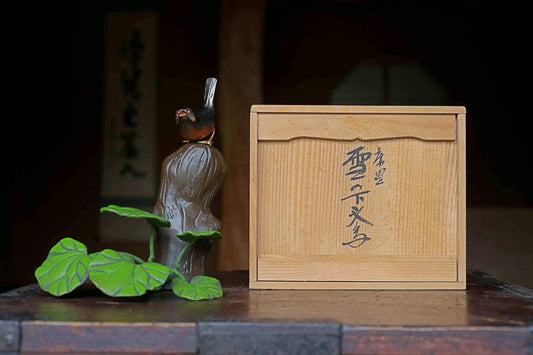A small figurine of a bird perched on a tree trunk, with a wooden box featuring Japanese characters next to it, both placed on a wooden surface.
