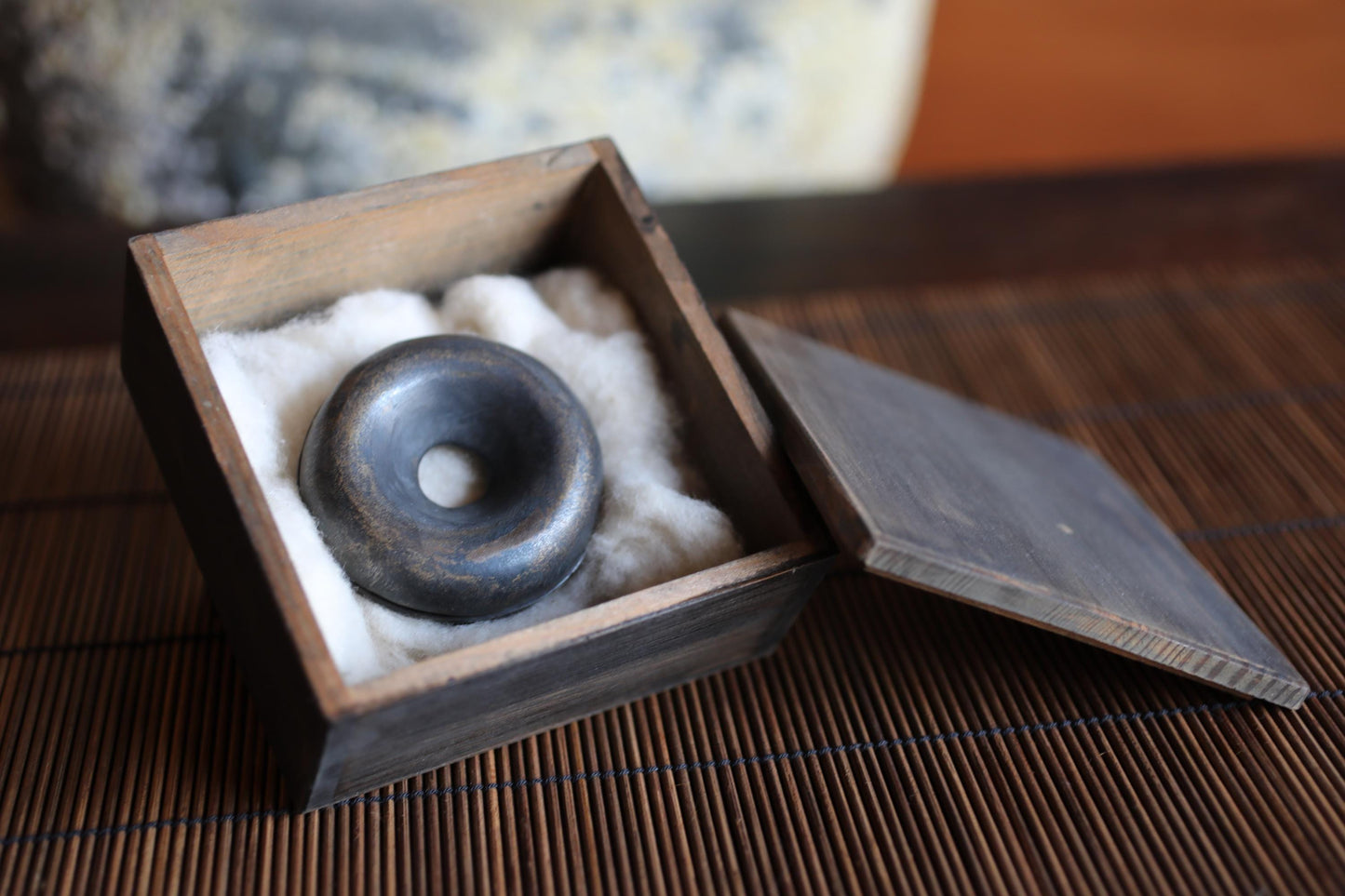 A wooden box containing a black metal object, possibly a piece of pottery or a sculpture, placed on a bamboo mat.