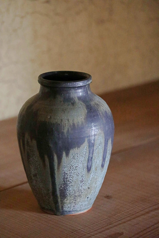 A ceramic vase with a blue and gray speckled pattern sits on a wooden surface against a plain wall.