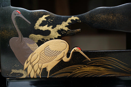 a black wooden surface with a decorative design of two large birds, possibly cranes, and a wave-like pattern. The birds are depicted in a stylized manner, with intricate details and patterns. The overall design is visually striking and artistic, showcasing the craftsmanship and creativity of the artist or designer.