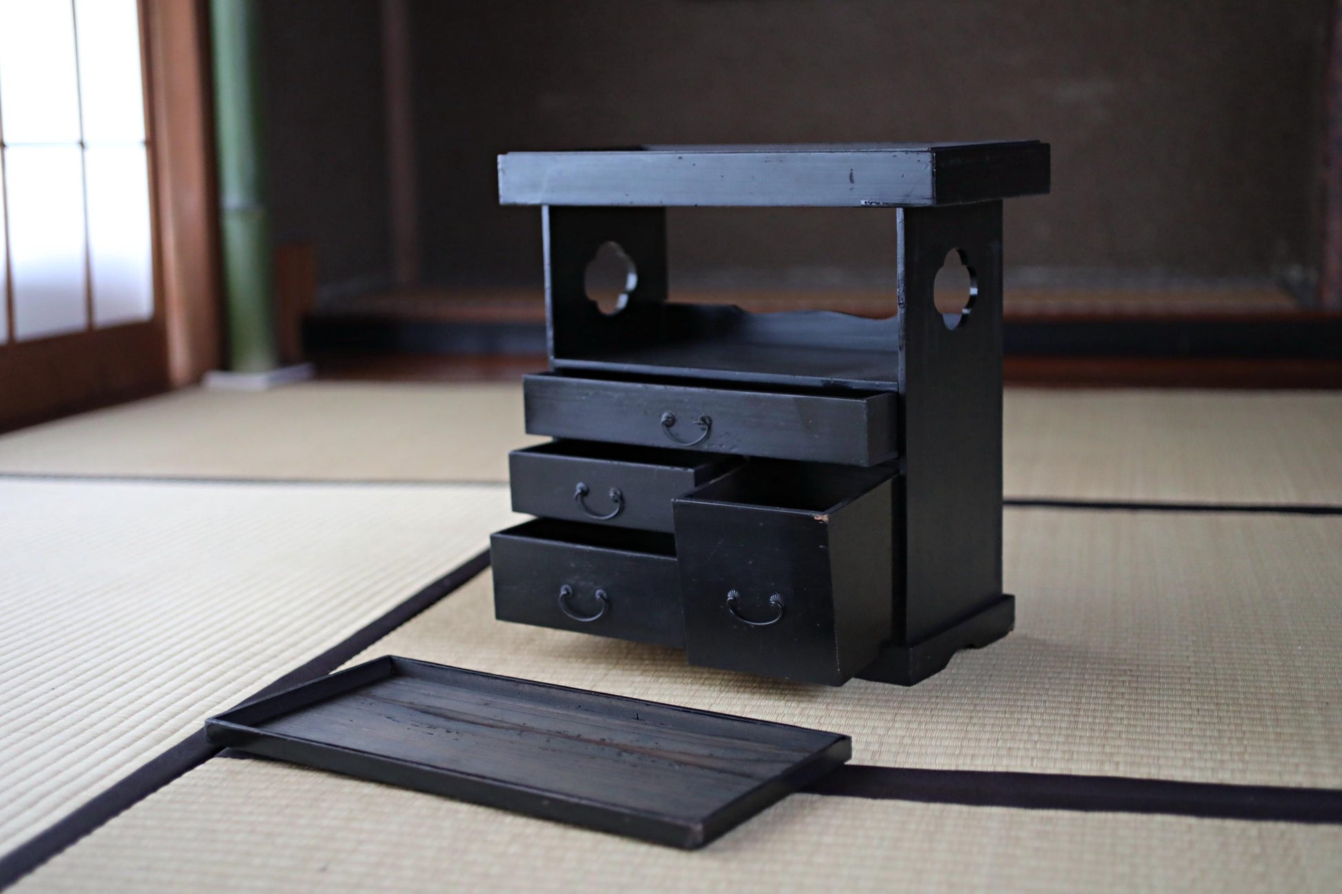 A black wooden cabinet with three drawers and a shelf is placed on a beige tatami mat in a room with a window.