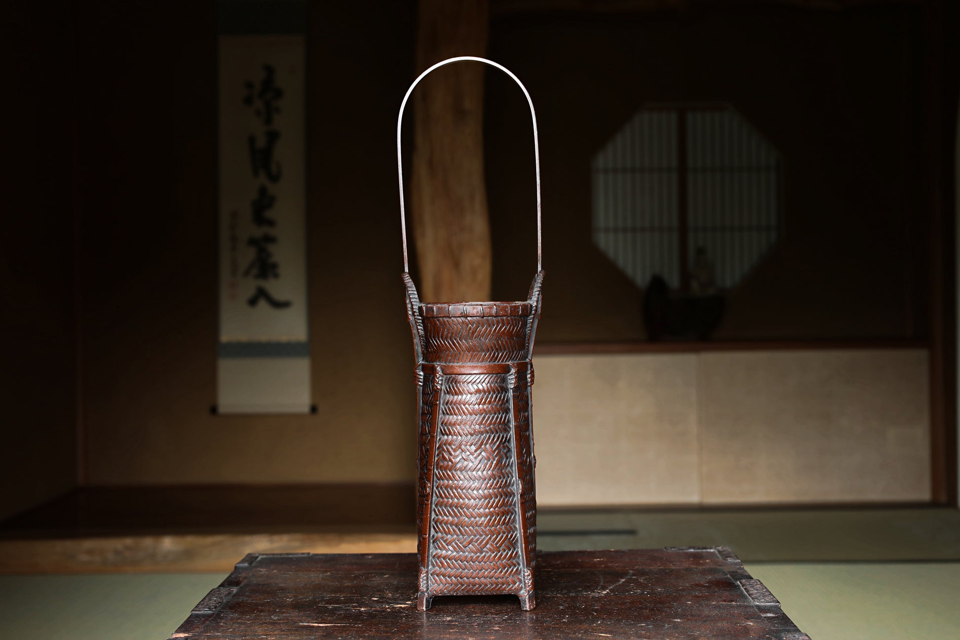 A traditional Japanese woven basket with a handle, placed on a wooden surface in a dimly lit room with a window and a sign with Japanese characters in the background.