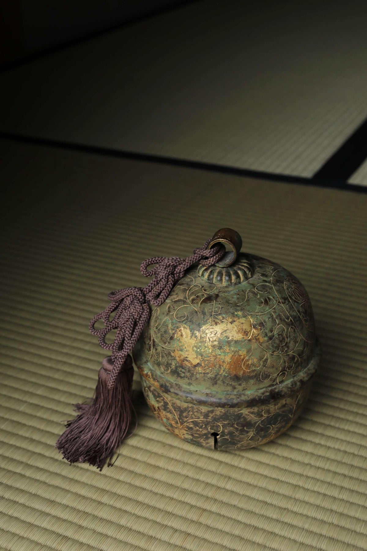 An ornate, antique-looking bell with a tassel hanging from it, resting on a woven mat.