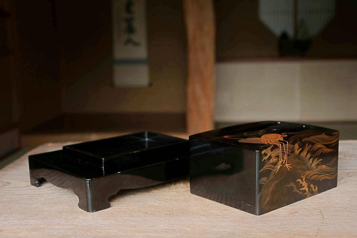 a wooden table with a black box and a decorative box placed on it. The decorative box has an intricate design with a bird and other elements.