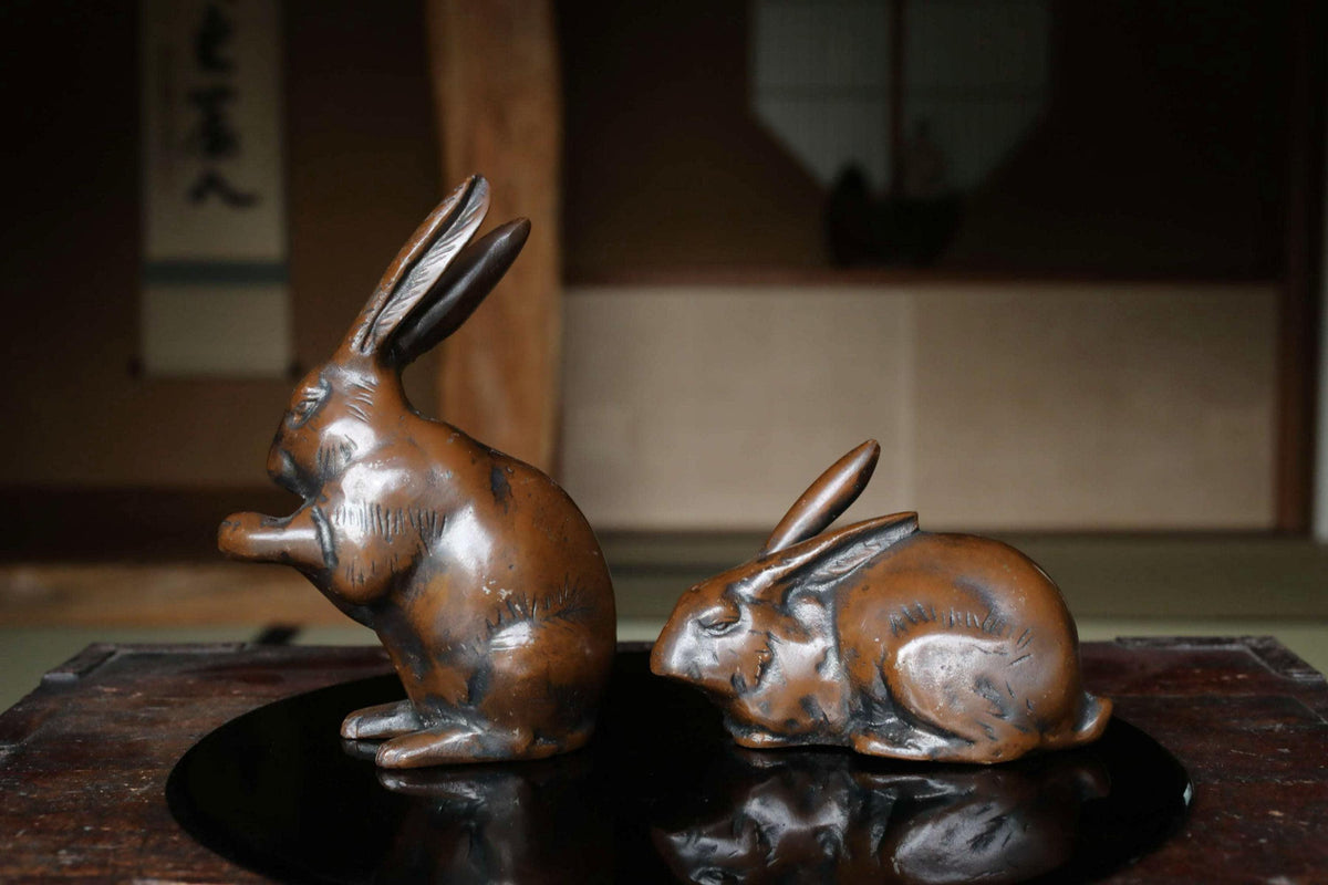 Two bronze rabbit figurines are displayed on a reflective surface, one standing and one sitting.