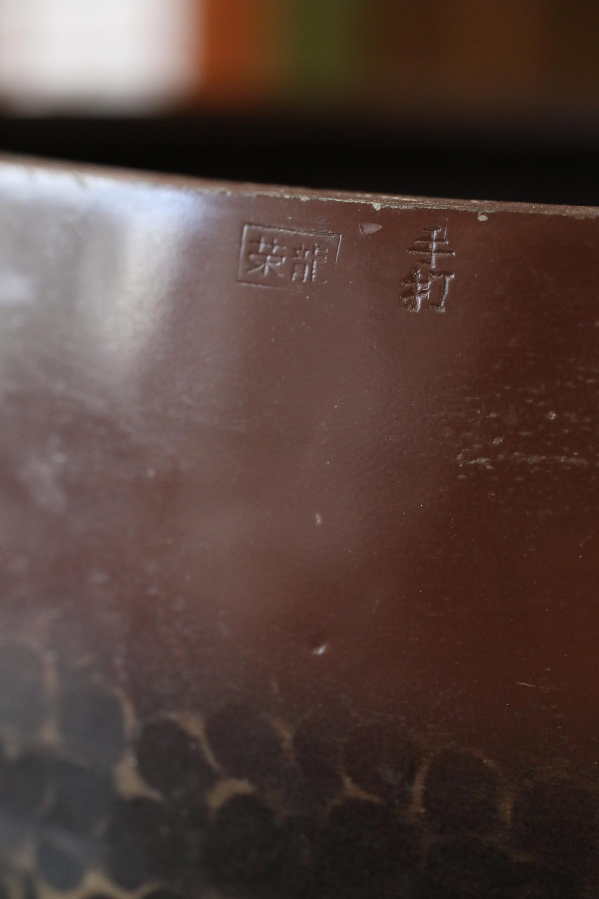 A close-up image of a brown, textured surface with a small, engraved symbol or text in the top left corner.