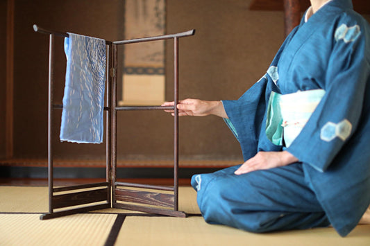 A person in a traditional Japanese kimono is sitting on the floor, reaching out to touch a wooden rack holding a blue cloth.
