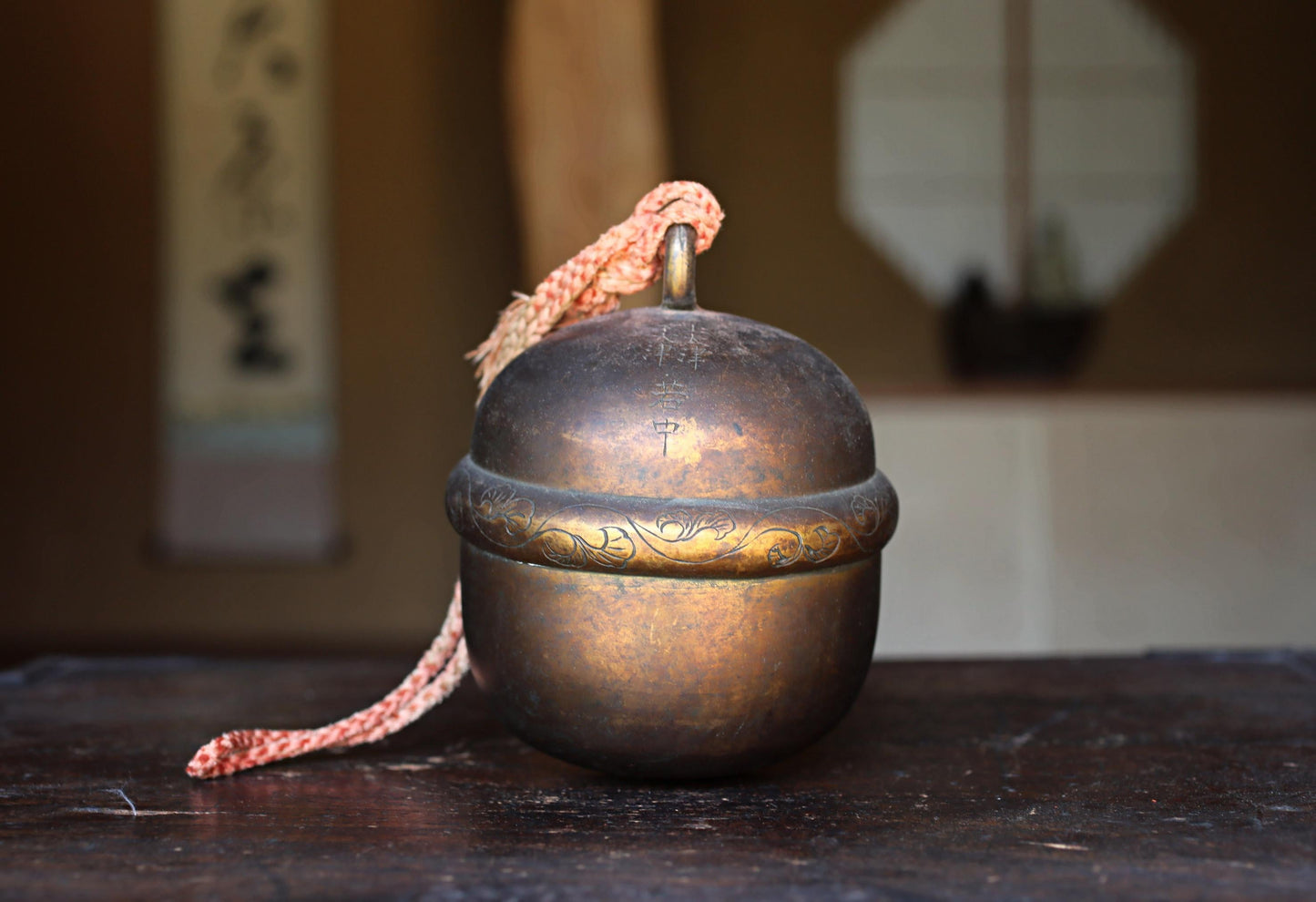 A brass bell with a decorative band sits on a wooden surface, with a blurred background that includes a window and a wall with Japanese characters.