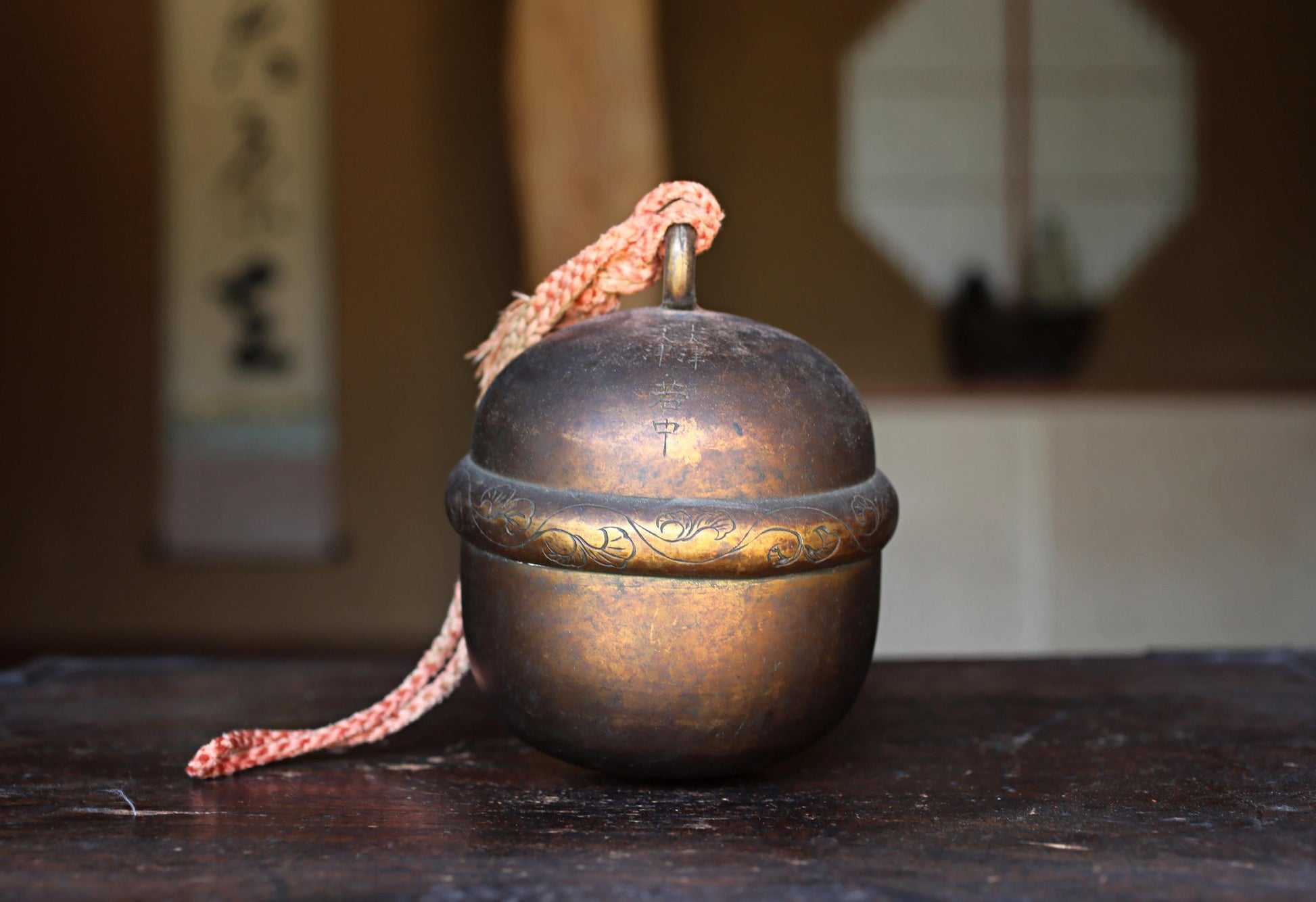 A brass bell with a decorative band sits on a wooden surface, with a blurred background that includes a window and a wall with Japanese characters.
