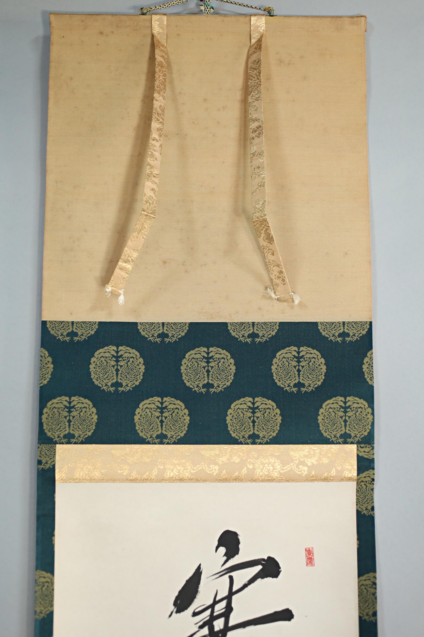 a wall hanging with a blue and gold pattern, a white paper with a black calligraphy design, and a gold ribbon hanging from the top of the wall hanging.