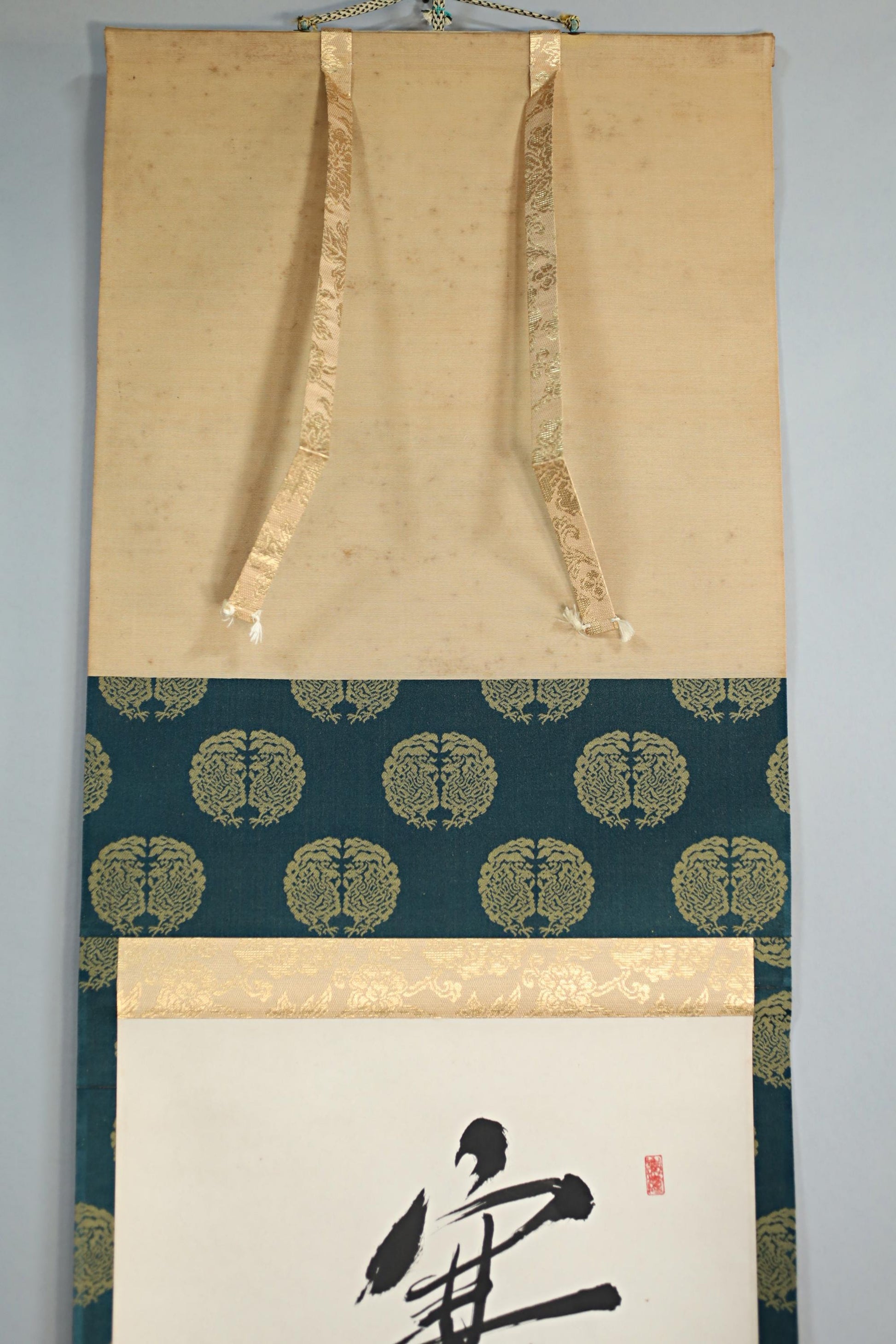a wall hanging with a blue and gold pattern, a white paper with a black calligraphy design, and a gold ribbon hanging from the top of the wall hanging.