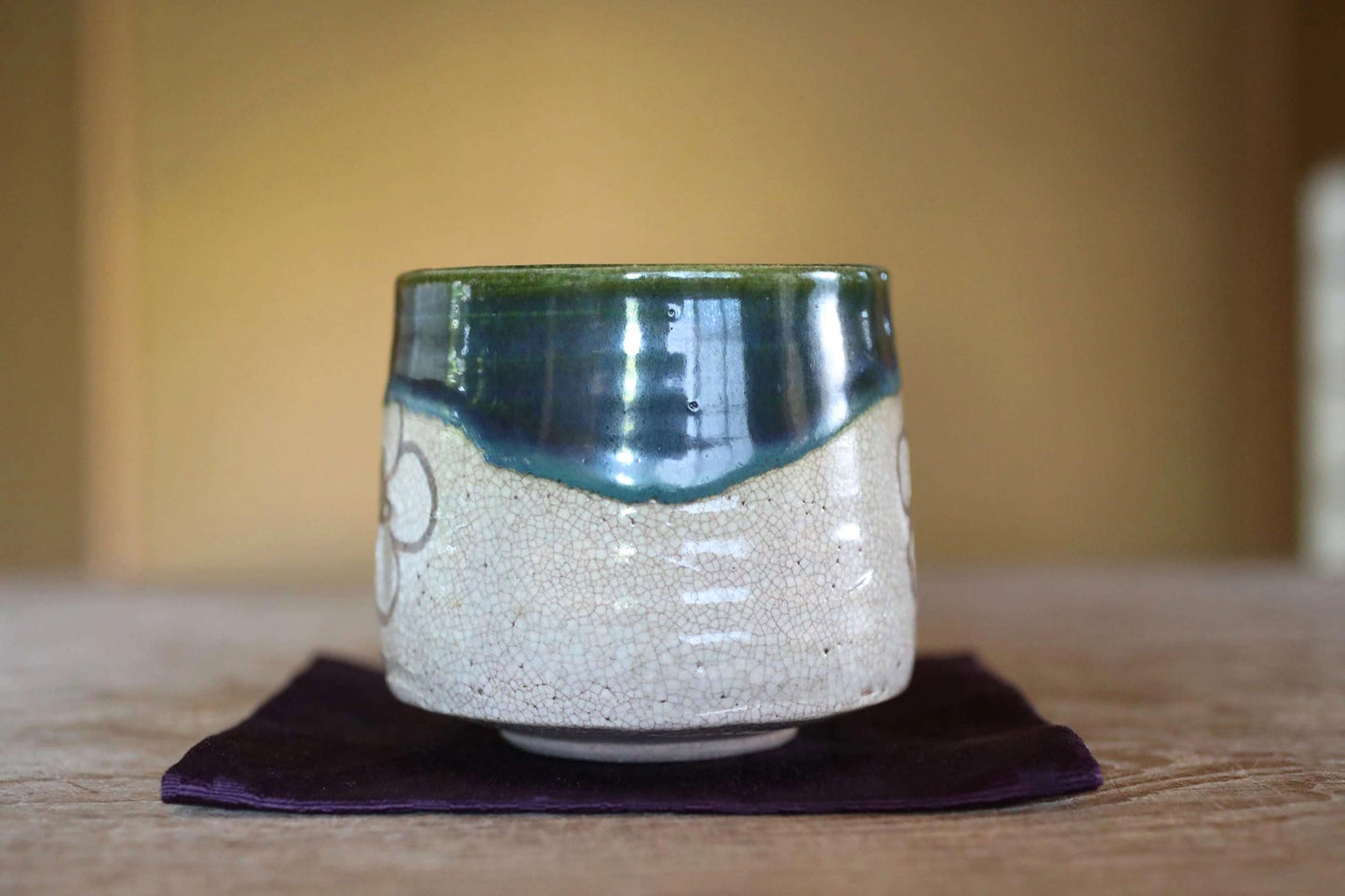 A ceramic mug with a green and white pattern sits on a purple cloth on a wooden surface.