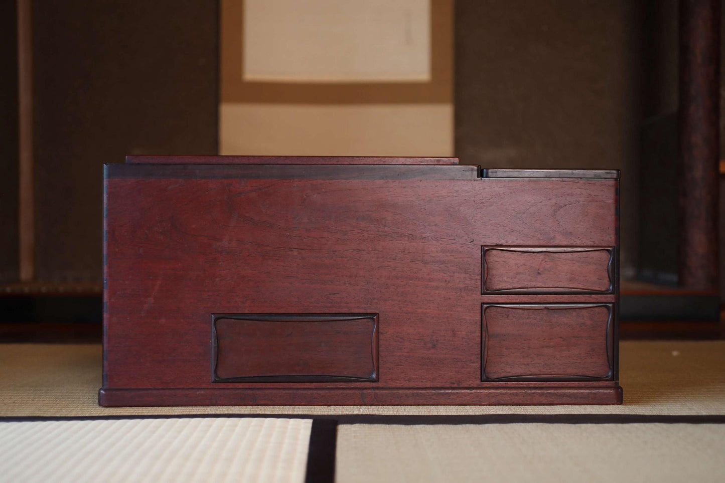 A wooden desk with two drawers and a shelf is placed on a tatami mat in a room with a wooden wall and a window.