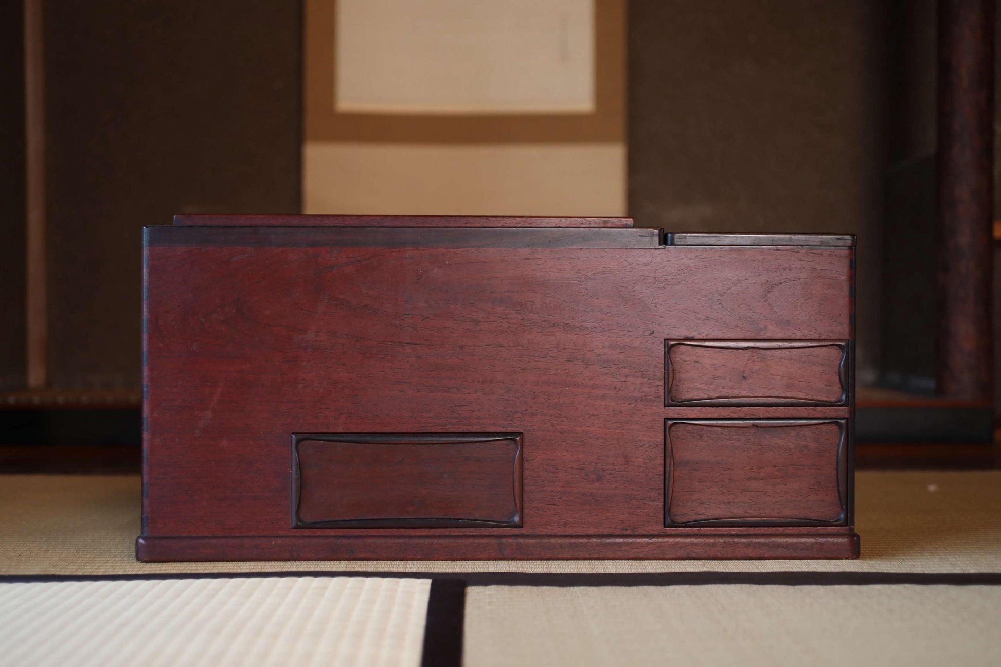 A wooden desk with two drawers and a shelf is placed on a tatami mat in a room with a wooden wall and a window.