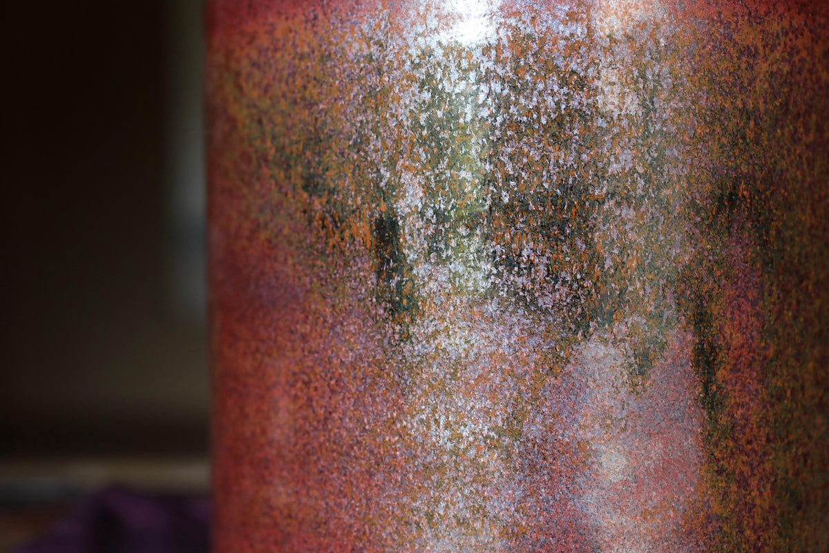 a close-up view of a textured surface with a reddish-brown hue, featuring a pattern of small dots or specks in various shades of orange, green, and brown.