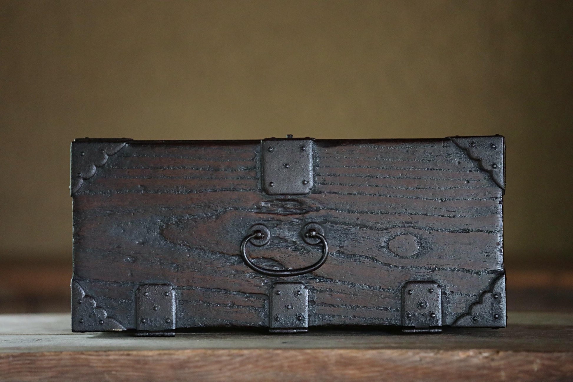 A dark wooden chest with metal hardware, including a handle and clamps, sits on a wooden surface.