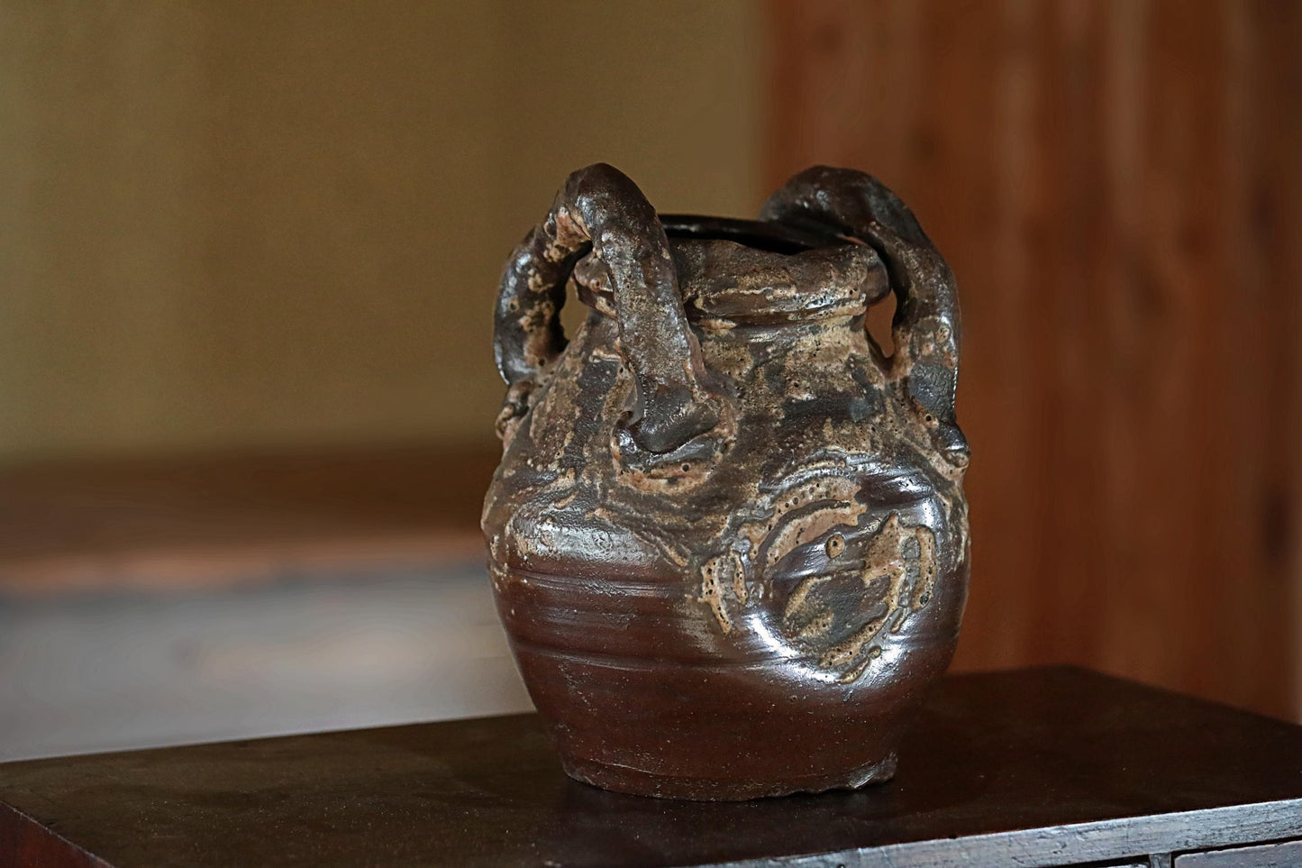 A dark brown, textured ceramic vase with a handle on top, sitting on a wooden surface.