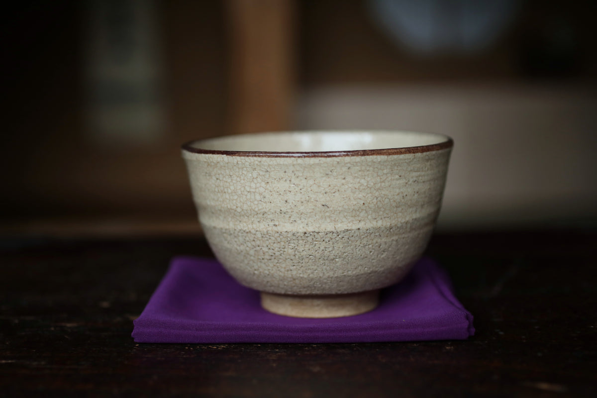 A white ceramic bowl with a brown rim sits on a purple cloth on a wooden surface.