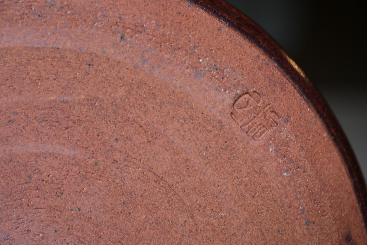 A close-up image of a red clay pot or vase, with a small engraved mark or logo on the surface.