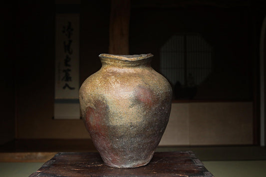 An old, weathered ceramic vase sits on a wooden surface, with a dark background that includes some text and a window.