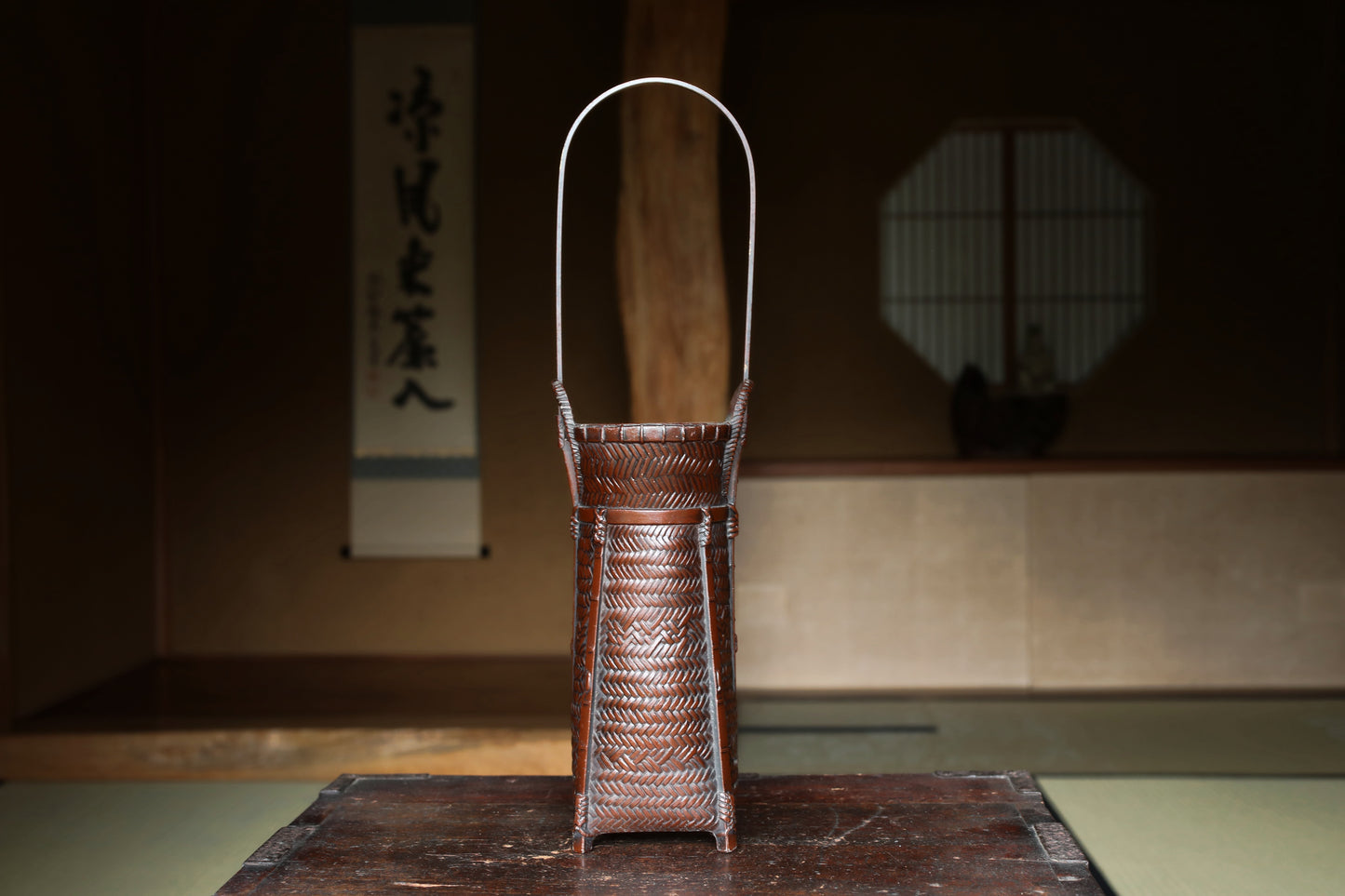 A traditional Japanese woven basket with a handle, placed on a wooden surface in a dimly lit room with a window and a wall with Japanese characters.