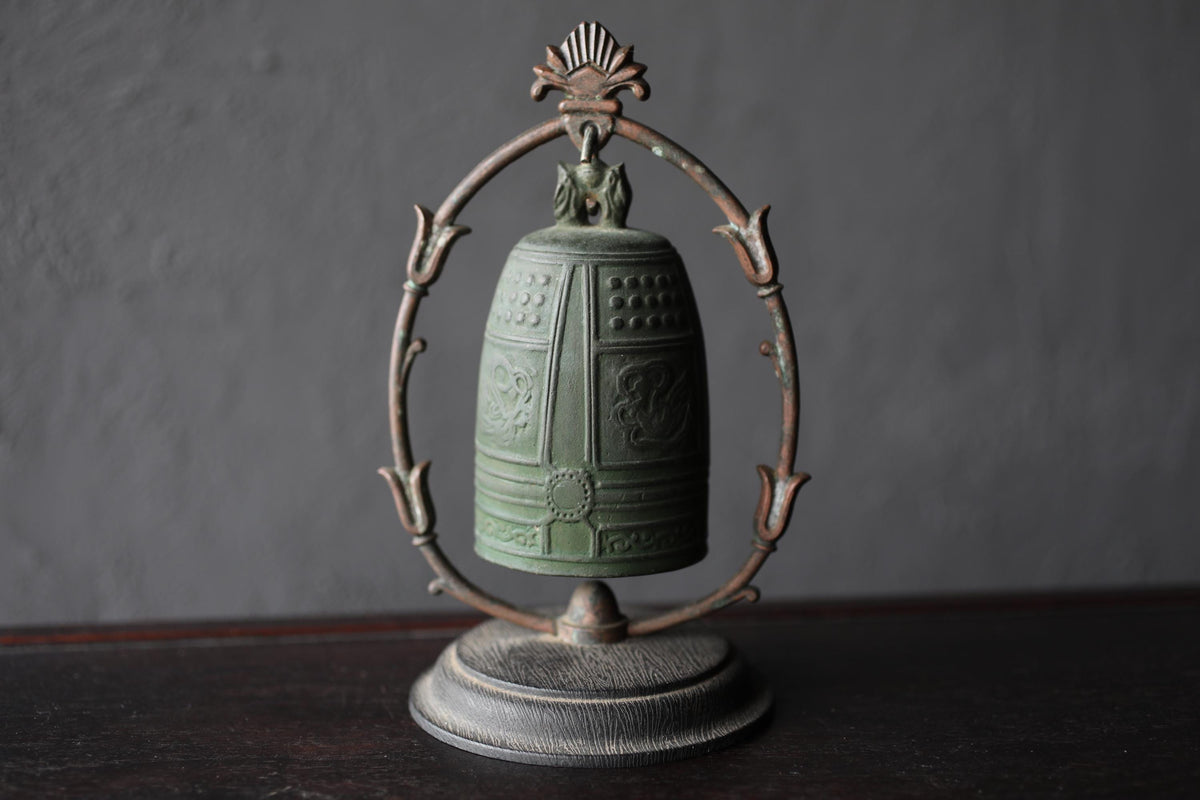 An antique green metal bell with a decorative metal frame, displayed on a dark wooden surface against a gray background.