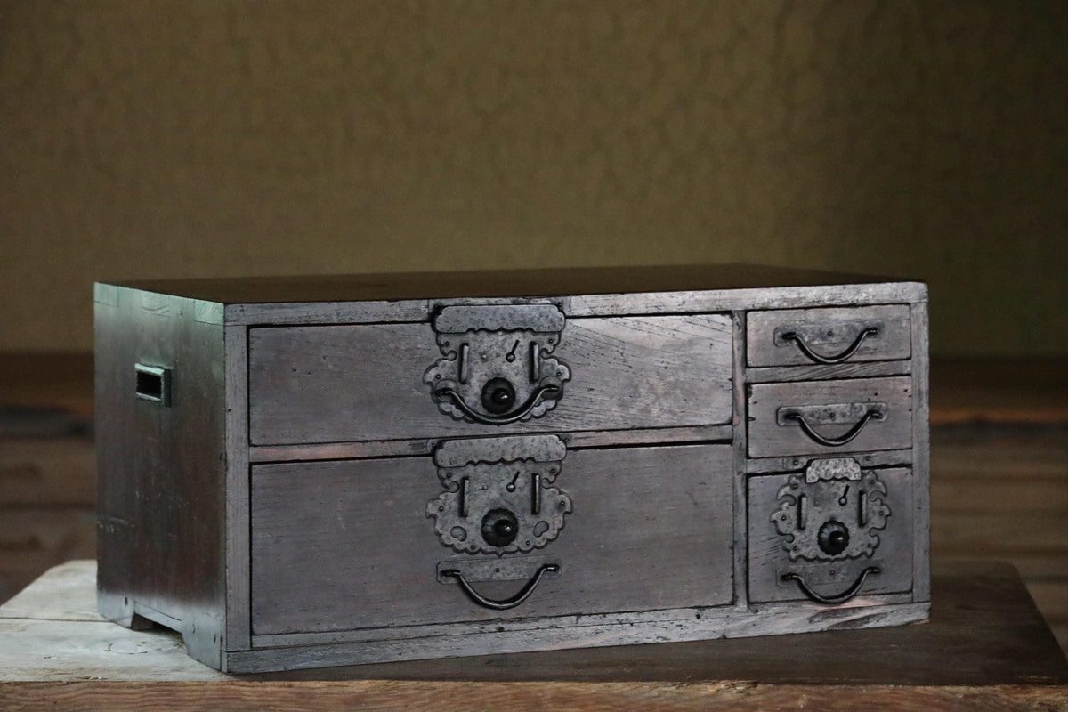 A wooden chest with six drawers, featuring intricate metal handles and decorations, sits on a wooden surface.