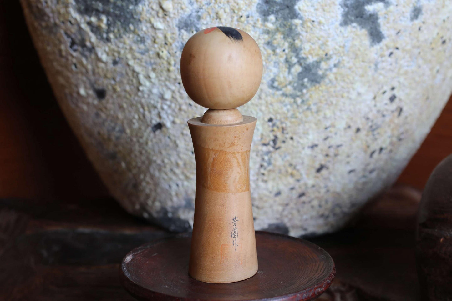 A wooden object with a round top and a textured body, placed on a wooden stand in front of a large, textured stone or ceramic piece.